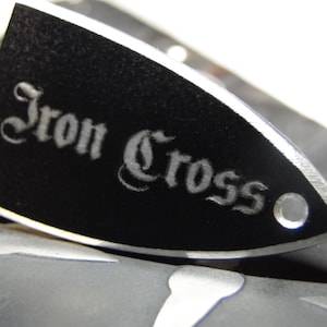 Op de afbeelding: Zwarte gitaarplectrum met een zilveren rand en de woorden "Iron Cross" in een zilveren, gestileerde lettertype.