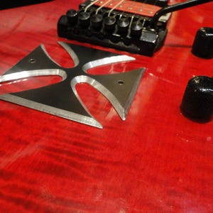 Puede incluir: Una guitarra el&eacute;ctrica roja con una incrustaci&oacute;n de cruz de metal negro en el cuerpo. La guitarra tiene botones negros y un puente negro.
