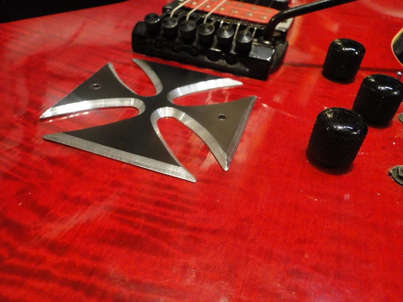 Puede incluir: Una guitarra el&eacute;ctrica roja con una incrustaci&oacute;n de cruz de metal negro en el cuerpo. La guitarra tiene botones negros y un puente negro.