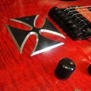 Puede incluir: Una guitarra el&eacute;ctrica roja con una incrustaci&oacute;n en forma de cruz negra en el cuerpo. La guitarra tiene un puente negro con clavijas de afinaci&oacute;n plateadas y perillas negras.