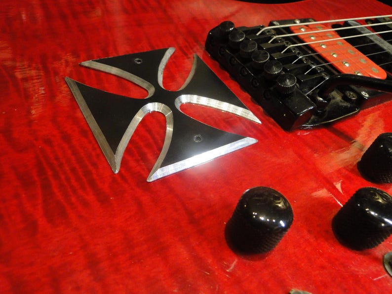 Puede incluir: Una guitarra el&eacute;ctrica roja con una incrustaci&oacute;n en forma de cruz negra en el cuerpo. La guitarra tiene un puente negro con clavijas de afinaci&oacute;n plateadas y perillas negras.