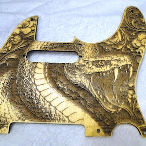 以下が含まれることがあります： 木製のギターピックガード。彫刻されたコブラのデザインが特徴です。蛇の頭部が大きく、牙がむき出しになっています。ピックガードは自然な木の色で、彫刻を際立たせるために濃い影がつけられています。