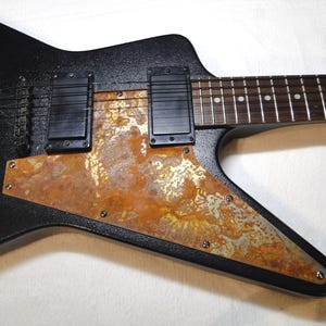 Könnte beinhalten: Schwarze E-Gitarre mit einem rostigen Metall-Pickguard. Die Gitarre hat einen schwarzen Korpus und einen schwarzen Kopf. Der Pickguard ist eine große, rostige Metallplatte, die den größten Teil des Gitarrenkörpers bedeckt.