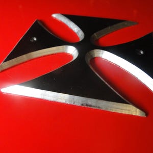 Puede incluir: Una cruz de metal negro con acabado plateado. La cruz tiene cuatro puntas y est&aacute; ligeramente elevada de la superficie roja sobre la que se encuentra.