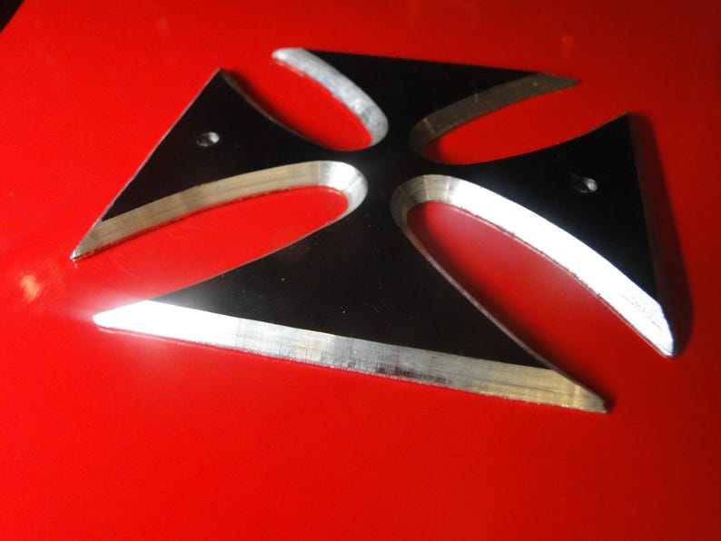 Puede incluir: Una cruz de metal negro con acabado plateado. La cruz tiene cuatro puntas y est&aacute; ligeramente elevada de la superficie roja sobre la que se encuentra.