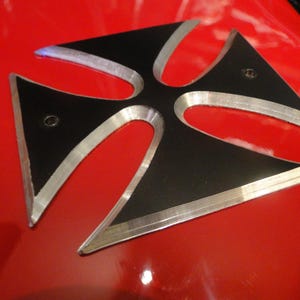 Puede incluir: Una cruz de metal negro y plateado con un borde elevado. La cruz est&aacute; montada sobre una superficie roja.