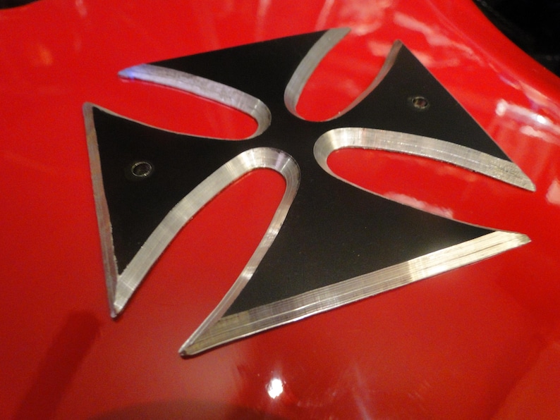 Puede incluir: Una cruz de metal negro y plateado con un borde elevado. La cruz est&aacute; montada sobre una superficie roja.