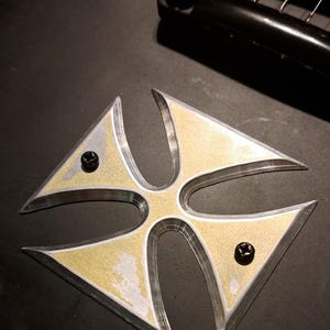 Può includere: Un'intarsio metallico a forma di croce argentata e dorata con due viti nere su un corpo di chitarra nero.
