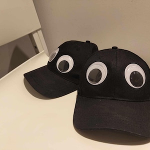 Può includere: Due cappellini da baseball neri con occhietti bianchi googly attaccati. I cappellini hanno una visiera curva e una parte posteriore in rete.