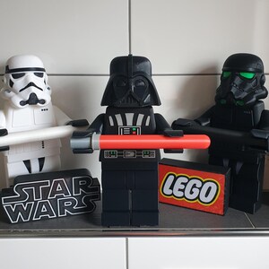 Könnte beinhalten: Drei schwarz-weiße Star Wars Lego-Figuren, Darth Vader, ein Stormtrooper und ein TIE-Fighter-Pilot. Die Figuren halten ihre charakteristischen Waffen, ein Lichtschwert, einen Blaster und eine Blaster-Pistole. Die Figuren stehen auf einem schwarzen Sockel mit den Worten "Star Wars" und "Lego" darauf.