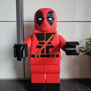 Könnte beinhalten: Eine rote Deadpool-Figur im Lego-Stil. Die Figur hat eine strukturierte rote Maske mit schwarzen Augenausschnitten, ein schwarzes Brustdesign und einen braunen Gürtel. Sie hält ein silbernes Schwert und hat schwarze Armaufsätze.