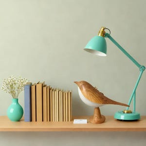 May include: A wooden bird sculpture with a white belly and brown wings sits on a wooden base. A teal blue desk lamp with a white shade is on a wooden shelf behind the bird. A stack of books is on the shelf to the left of the bird.