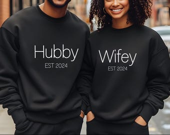May include: Matching black sweatshirts with white text that reads "Hubby EST 2024" and "Wifey EST 2024". The sweatshirts are a solid black color and have a crew neck design. The image shows two people wearing the sweatshirts.