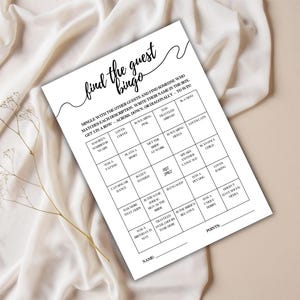 May include: A white "Find the Guest Bingo" game sheet with black text, featuring a grid of descriptive prompts. The sheet is on a beige surface with a sprig of dried flowers.