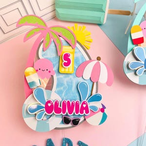May include: A colorful papercraft pool party decoration featuring a pool, palm tree, ice cream, and the name "Olivia". Includes a sun, sunglasses, and a beach ball. A turquoise popsicle is also visible.