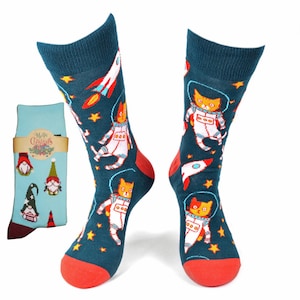 May include: A pair of teal blue socks with a pattern of orange cats in astronaut suits and red rockets on a dark blue background with white stars. The socks have red and white trim at the top and red trim at the bottom.