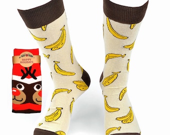 May include: A pair of cream-colored socks with a brown cuff and a yellow banana pattern. The socks are packaged with a red and white sock with a reindeer face and the text "Happy Holidays".
