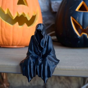 May include: A black 3D printed figurine of a hooded figure sitting with its head down. The figurine is in front of two jack-o'-lanterns.