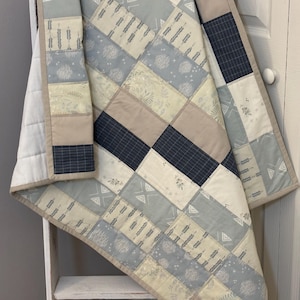 May include: A patchwork quilt featuring rectangular fabric panels in shades of navy blue, light blue, cream, and beige. The design includes various patterns such as thin grid lines, botanical prints, and geometric shapes, finished with a beige border.