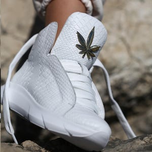 Puede incluir: Zapatillas blancas de caña alta con estampado de piel de serpiente y un diseño de hoja de marihuana dorada.