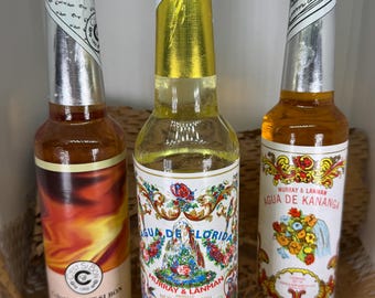May include: Three glass bottles of cologne. The left bottle is labeled C'est Si Bon Pusanga with a fiery orange and red graphic. The center bottle is labeled Agua de Florida Murray & Lanman with a colorful floral and mountain illustration. The right bottle is labeled Agua de Kananga Murray & Lanman with a floral bouquet graphic.