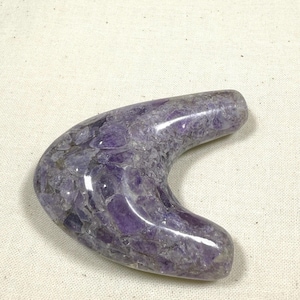 May include: A boomerang-shaped massage tool crafted from polished amethyst stone. The surface features a mosaic of deep purple and translucent violet crystal fragments embedded within a smooth, glossy, and rounded structure.