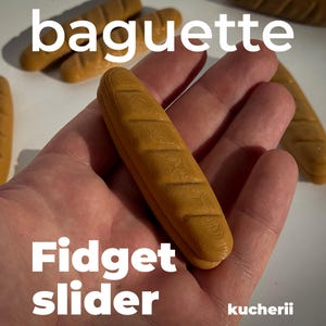 Op de afbeelding: Een bruine, baguette-vormige fidget slider wordt in een hand gehouden. Het woord "baguette" staat in het wit boven de slider en "Fidget slider" staat eronder. Het woord "kucherii" staat rechtsonder.