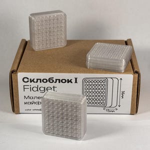 Puede incluir: Se muestran tres juguetes fidget cuadrados translúcidos con una superficie texturizada. Los juguetes están sobre y alrededor de una caja de cartón etiquetada como "Склоблок I Fidget". La caja también muestra un diagrama con dimensiones de 1,3 cm x 3,6 cm.