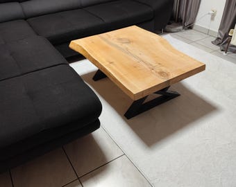 May include: A light-colored wooden coffee table with a live edge design. The table sits on a black metal base. A black sectional sofa is in the background. The table is approximately 40 inches long and 20 inches wide.