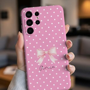 Puede incluir: Funda de teléfono rosa con lunares blancos, un lazo rosa claro y el nombre "Annie" en cursiva. La funda tiene múltiples recortes para la cámara y se sostiene en una mano.