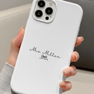 May include: White phone case with a black camera module and the text "Mrs. Miller" in a handwritten font, with a small bow graphic. The case is held in a person's hand.
