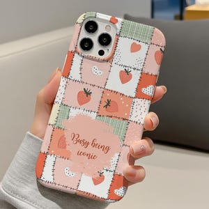 Puede incluir: Una funda de teléfono con un diseño de patchwork con fresas, corazones y lunares en tonos rosa, rojo y verde. La funda tiene el texto "Busy being iconic" sobre un fondo rosa.