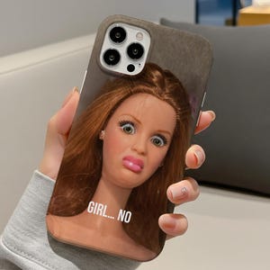 May include: A phone case featuring a doll's face with wide eyes and a surprised expression. The doll has long auburn hair and pink lips. The text "GIRL... NO" is printed on the case.