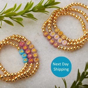 May include: Three gold beaded bracelets with colorful letter beads spelling out the names Avery, Grace, and Zoe. The bracelets are on a white surface.