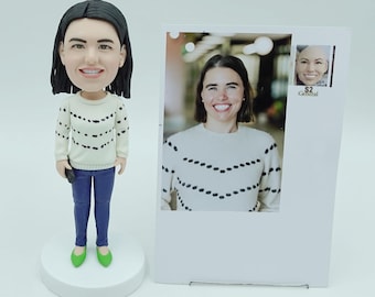 May include: A custom bobblehead figurine of a person wearing a white sweater with black dot patterns, blue jeans, and green shoes. The figurine is standing on a white base next to a photo of the same person wearing a similar sweater. A small stamp is visible on the photo.