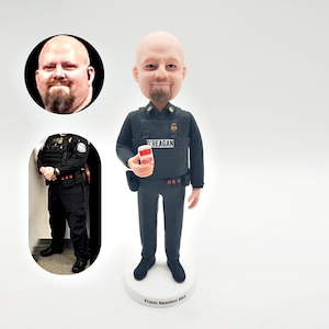 May include: A bobblehead figurine of a man in a police uniform holding a small carton of milk. The figurine has a bald head and a goatee. The uniform includes a vest with the name "REAGAN" on it. The base of the figurine reads "From Session 664".