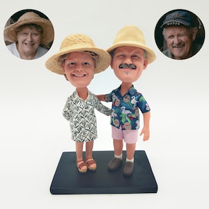 May include: A custom bobblehead figurine of a couple, both wearing straw hats. The male figure has a moustache and is wearing a Hawaiian shirt and pink shorts. The female figure is wearing a patterned dress. The base is black.