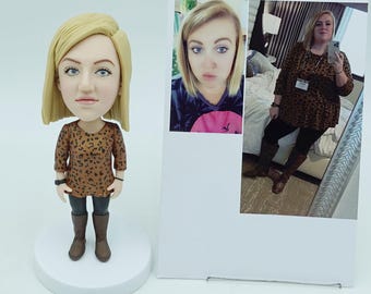May include: A bobblehead figurine of a woman with blonde hair, wearing a brown leopard print top, gray pants, and brown boots. The figurine is on a white base. A photo of the woman is next to the figurine.
