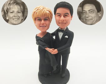 May include: A figurine of a couple in formal attire, with a woman in a black dress and a man in a tuxedo. The figurine is on a black base, with two circular portraits in the background. The woman has blonde hair and the man has black hair.