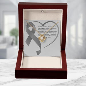 May include: A gold-coloured knot necklace presented in an open, dark wooden box. The box displays the necklace on a card with a grey ribbon and text. The text reads, "In Celtic tradition, the knot has no beginning and no end..."