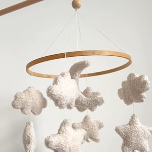 May include: A baby mobile with soft, plush clouds, stars, and a crescent moon in off-white. The wooden frame and hanging arm add a natural touch. This nursery decor item is designed to gently rotate.
