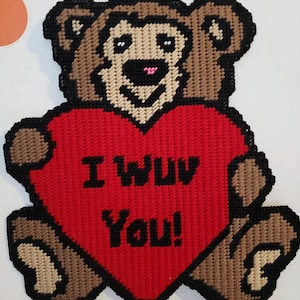 May include: A handmade, wall-hanging teddy bear made from plastic canvas. The bear is brown, tan, and black, holding a red heart with the words "I WUV You!" in black. The bear has a pink nose and is hanging from a black string.
