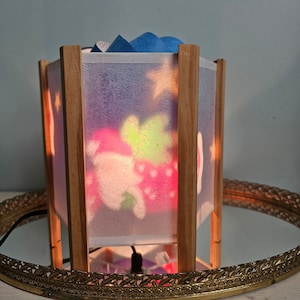 May include: A white, wooden, hexagonal lampshade with a light blue fabric lining. The lampshade has a colorful, cartoon-like design on the front, featuring a pink and green character with a red heart. The lampshade is sitting on a gold, decorative tray.