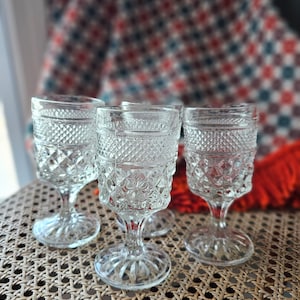 Puede incluir: Cuatro vasos de cristal transparente con tallo y un patrón de diamante. Los vasos están sobre una superficie de mimbre tejido.