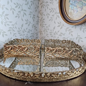Op de afbeelding: Een ovale dienblad van verguld metaal met een gespiegelde oppervlak en een decoratieve filigraanrand. Twee kleinere, rechthoekige, opengewerkte dienbladen met bloemmotieven staan op het grotere dienblad.