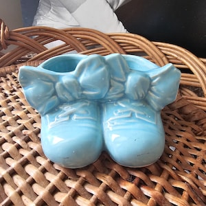 Pode incluir: Um vaso de cerâmica azul claro em forma de sapatos com laços no topo. O vaso está sobre uma superfície de vime trançado.