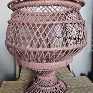 Puede incluir: Un macetero de mimbre rosa con una base redonda y una parte superior ancha y abierta. El macetero está hecho de mimbre tejido y tiene un diseño delicado y femenino.