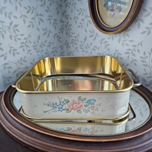 Puede incluir: Una fuente rectangular de metal blanquecino con un diseño floral de rosas rosas y nomeolvides azules. La fuente tiene un borde y asas doradas. Se encuentra sobre un espejo redondo con marco marrón.