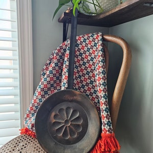 May include: A vintage copper or brass wall hanging with a floral design. The hanging is attached to a black metal hook and is hanging on a chair with a red and white plaid throw blanket.