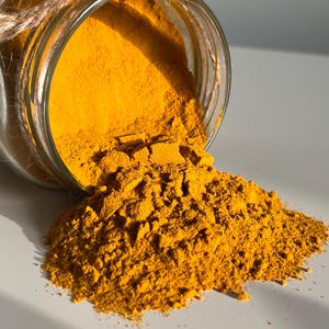 Op de afbeelding: Een heldere glazen pot met een jute touw om de rand, gevuld met felgeel kurkumapoeder. De pot is gekanteld, waardoor het poeder op een wit oppervlak wordt gemorst. Het poeder is fijn gemalen en heeft een levendige kleur.